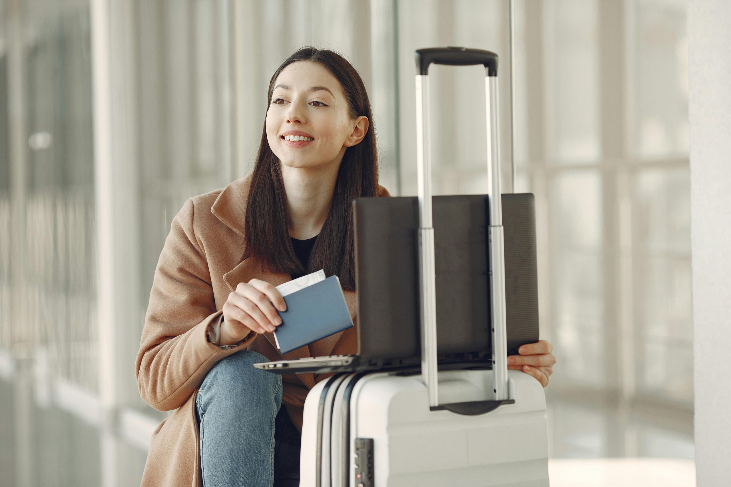 A woman sitting with her laptop and luggage, holding a passport, waiting in an airport terminal.